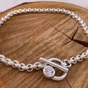 Könnte beinhalten: Eine silberne Kettenkette mit einem Toggle-Verschluss mit einem klaren Kristall. Die Kette besteht aus kleinen, runden Gliedern.