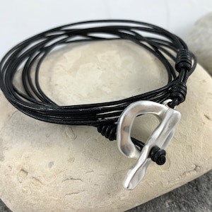 Op de afbeelding: Een zwarte leren armband met een zilveren sluiting. De armband is gelaagd en heeft een geknoopt detail.