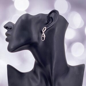 May include: Silver-toned dangle earrings with a black oval stone and a small, clear accent stone. The earrings are displayed on a black mannequin head against a blurred, light-colored background.