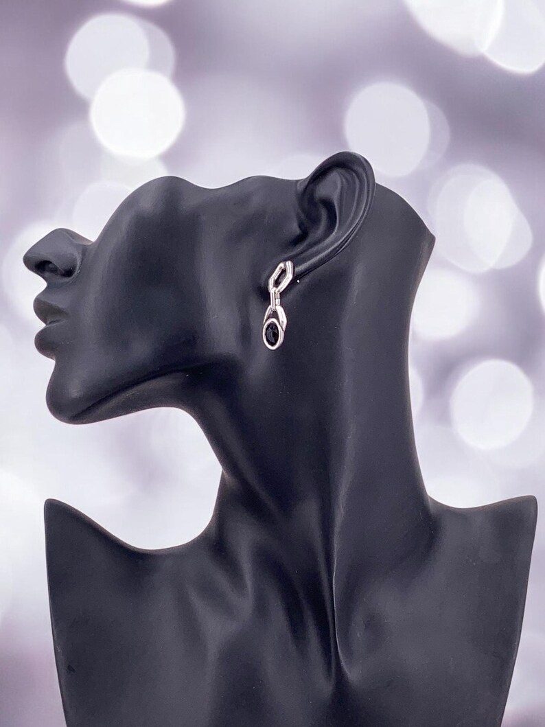 May include: Silver-toned dangle earrings with a black oval stone and a small, clear accent stone. The earrings are displayed on a black mannequin head against a blurred, light-colored background.