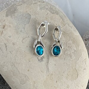 May include: A pair of silver-toned drop earrings. Each earring features a chain-link design with a teal-colored, oval-shaped gemstone at the bottom. The earrings are displayed on a light-colored, textured stone surface.