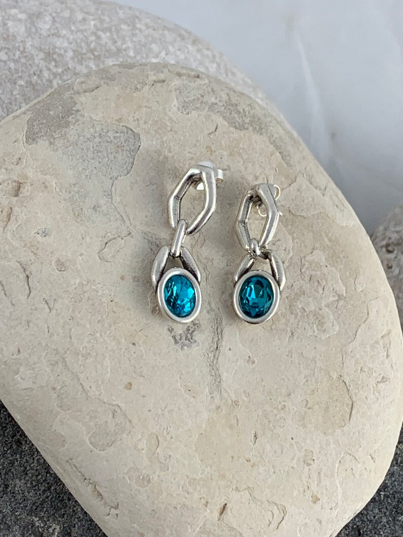May include: A pair of silver-toned drop earrings. Each earring features a chain-link design with a teal-colored, oval-shaped gemstone at the bottom. The earrings are displayed on a light-colored, textured stone surface.