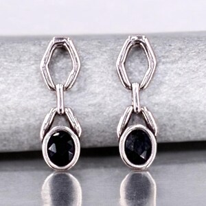 May include: A pair of silver-toned drop earrings. Each earring features a black oval-shaped gemstone suspended from a geometric link. The earrings are displayed on a light gray surface, showcasing their reflective qualities.