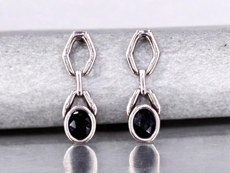 May include: A pair of silver-toned drop earrings. Each earring features a black oval-shaped gemstone suspended from a geometric link. The earrings are displayed on a light gray surface, showcasing their reflective qualities.