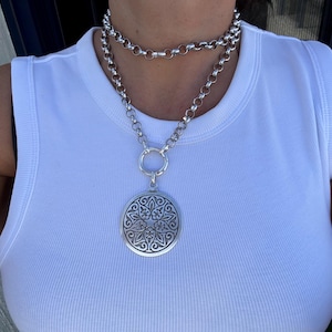 May include: A silver-toned necklace with a large, round pendant featuring an intricate floral design. The necklace has a double-strand chain and a large ring clasp. The pendant hangs from a shorter chain.