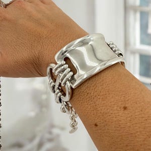 Può includere: Un braccialetto d'argento con un grande ciondolo rettangolare piatto e una catena fatta di cerchi collegati. Il braccialetto è al polso di una persona.