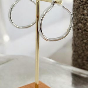 Puede incluir: Pendientes de aro plateados exhibidos en un soporte dorado con base de madera. Los pendientes tienen un dise&ntilde;o redondeado y aplanado. El soporte se encuentra sobre un fondo neutro, resaltando las joyas.