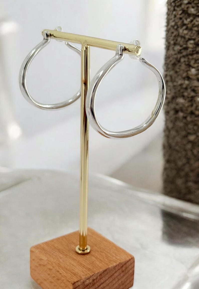 Puede incluir: Pendientes de aro plateados exhibidos en un soporte dorado con base de madera. Los pendientes tienen un dise&ntilde;o redondeado y aplanado. El soporte se encuentra sobre un fondo neutro, resaltando las joyas.