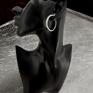 Puede incluir: Un pendiente de aro plateado se muestra en una cabeza de maniqu&iacute; negra. El pendiente tiene un dise&ntilde;o simple y pulido, con una forma suave y redondeada. El maniqu&iacute; se encuentra sobre un fondo plateado texturizado.