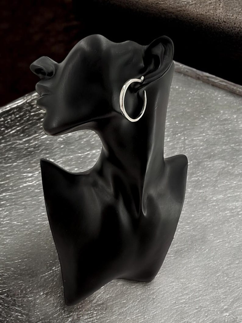 Puede incluir: Un pendiente de aro plateado se muestra en una cabeza de maniqu&iacute; negra. El pendiente tiene un dise&ntilde;o simple y pulido, con una forma suave y redondeada. El maniqu&iacute; se encuentra sobre un fondo plateado texturizado.