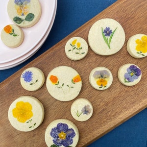 May include: A wooden board with a variety of round cookies decorated with pressed flowers. The cookies are decorated with yellow, orange, purple, and blue flowers.