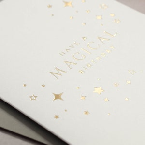 May include: White card with gold foil lettering that says "Have a Magical Birthday" with gold foil stars scattered around the text.