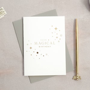 May include: A white card with gold foil lettering that reads "Have a Magical Birthday" with a design of gold stars in the shape of a crescent moon.