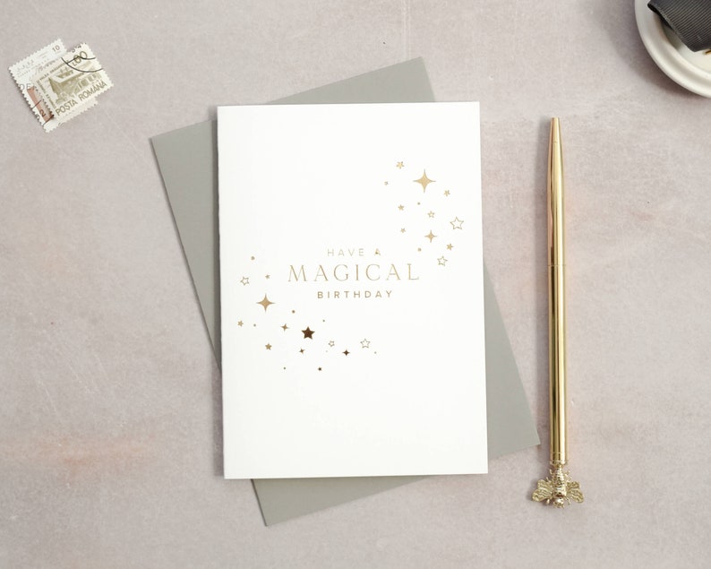 May include: A white card with gold foil lettering that reads "Have a Magical Birthday" with a design of gold stars in the shape of a crescent moon.