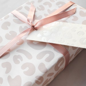 May include: A white gift box with a pink leopard print design. The box is wrapped with a pink satin ribbon and has a white gift tag attached.