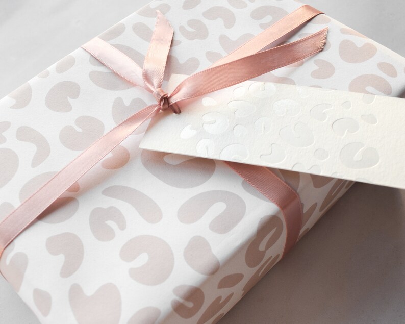 May include: A white gift box with a pink leopard print design. The box is wrapped with a pink satin ribbon and has a white gift tag attached.