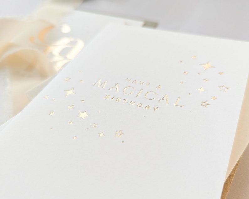 May include: A white card with gold foil lettering that reads "Have a Magical Birthday". The card is decorated with a scattering of gold foil stars.