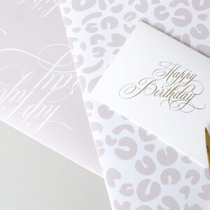 May include: A white card with gold lettering that says "Happy Birthday". The card is on top of a sheet of wrapping paper with a pink leopard print pattern. A pair of gold scissors is on the right side of the image.