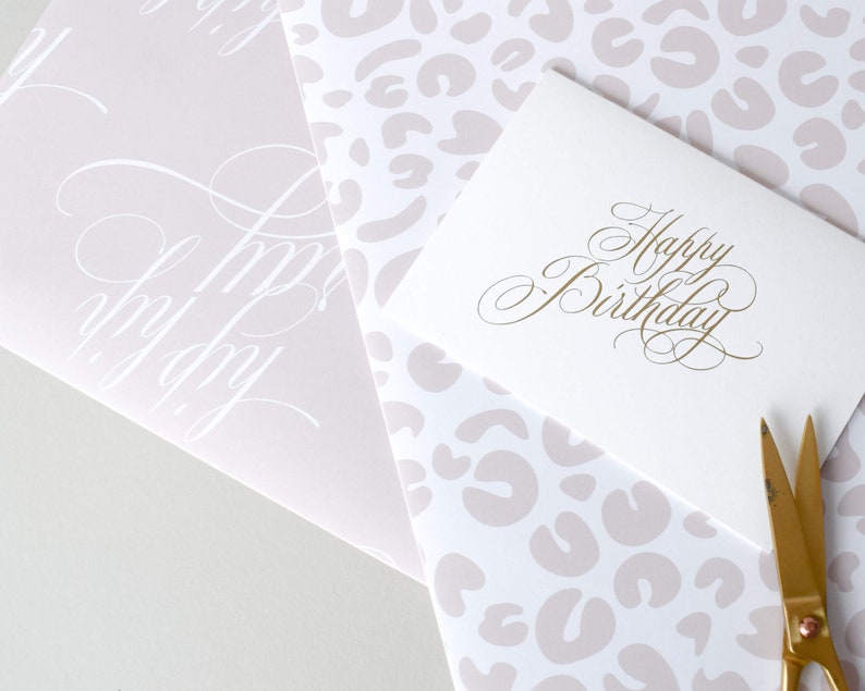 May include: A white card with gold lettering that says "Happy Birthday". The card is on top of a sheet of wrapping paper with a pink leopard print pattern. A pair of gold scissors is on the right side of the image.