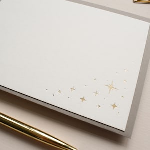May include: A rectangular white note card featuring a gold foil-stamped star pattern in the bottom right corner. The card is layered over a light gray envelope, and a gold metallic pen is positioned diagonally across the bottom left corner.