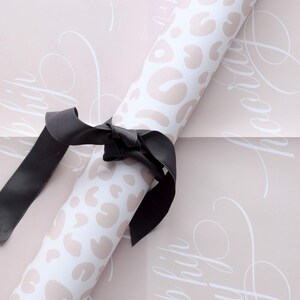 May include: A roll of pink wrapping paper with a white leopard print and the words "Happy" repeated in a cursive font. A black ribbon is tied around the roll.