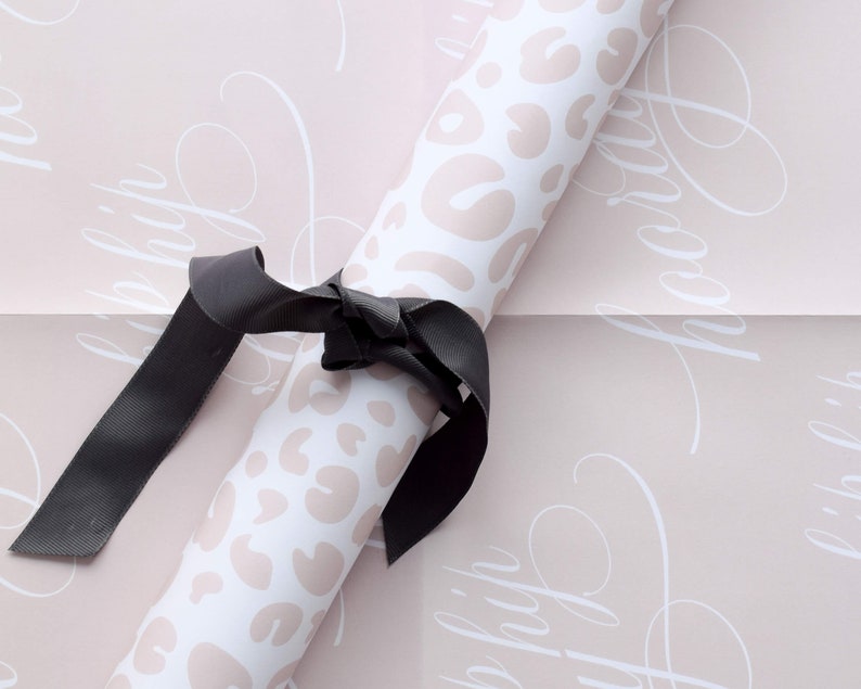 May include: A roll of pink wrapping paper with a white leopard print and the words "Happy" repeated in a cursive font. A black ribbon is tied around the roll.