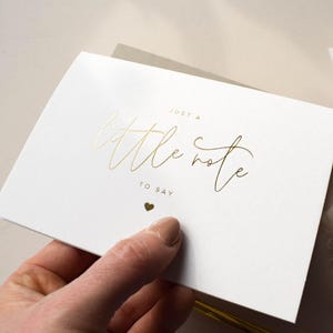 Pode incluir: Um cart&atilde;o branco com letras douradas que dizem "Just a little note to say" e um pequeno cora&ccedil;&atilde;o. O cart&atilde;o &eacute; segurado por uma pessoa.