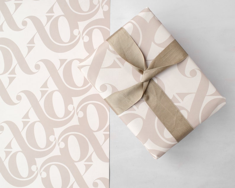 May include: A gift box wrapped in beige patterned paper with a brown satin ribbon tied in a bow. The paper has a repeating pattern of stylised letters.