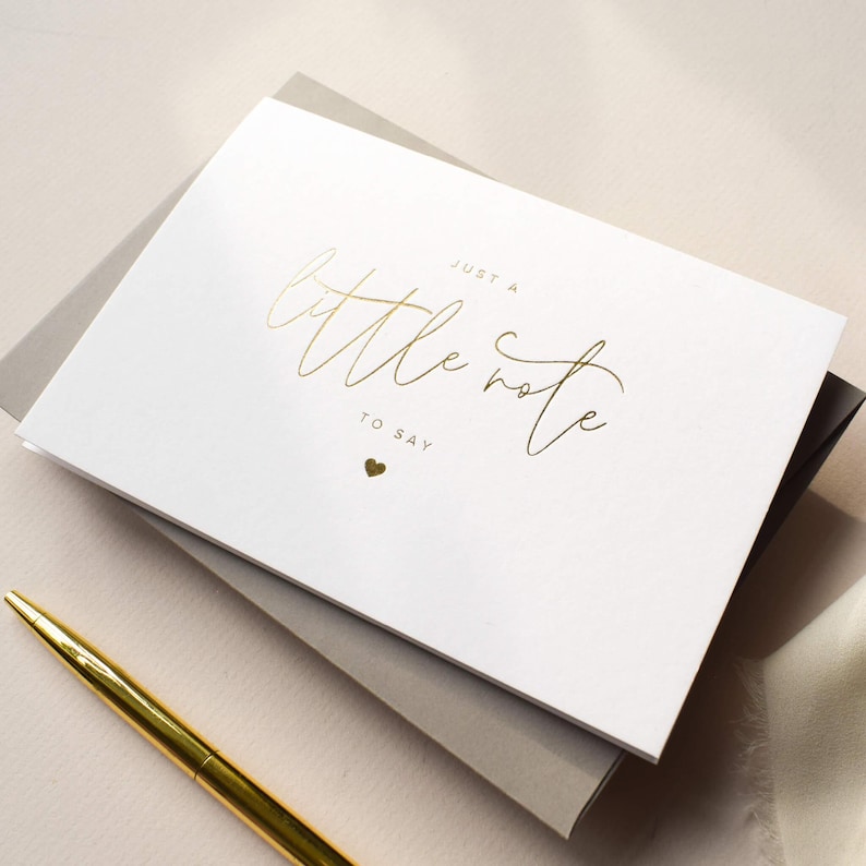 Pode incluir: Um cart&atilde;o branco com texto dourado em it&aacute;lico que diz "Just a little note to say" e um pequeno cora&ccedil;&atilde;o dourado. O cart&atilde;o est&aacute; em cima de um envelope cinza, com uma caneta dourada em primeiro plano.