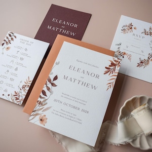 May include: A set of wedding stationery, including invitations and an order of the day, with the names "Eleanor" and "Matthew." The designs feature autumnal floral and leaf motifs in brown and orange hues. The stationery is displayed on a pale pink surface.