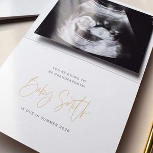 Pode incluir: Cart&atilde;o branco de an&uacute;ncio de gravidez com imagem de ultrassom e texto dourado. O texto diz "Voc&ecirc;s v&atilde;o ser av&oacute;s!" e "Baby Smith nasce no ver&atilde;o de 2026."