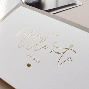 Pode incluir: Um cart&atilde;o branco com a frase dourada "Just a little note to say" e um pequeno cora&ccedil;&atilde;o dourado. O cart&atilde;o est&aacute; sobre um fundo cinza claro.