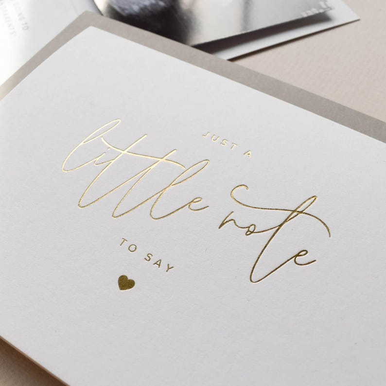 Pode incluir: Um cart&atilde;o branco com a frase dourada "Just a little note to say" e um pequeno cora&ccedil;&atilde;o dourado. O cart&atilde;o est&aacute; sobre um fundo cinza claro.