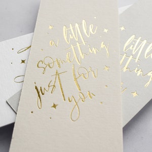 May include: Two white gift tags with gold foil lettering. The top tag reads "a little something just for you." The bottom tag reads "a little thing."