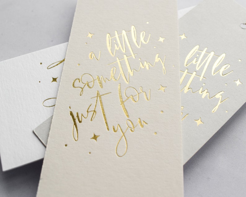 May include: Two white gift tags with gold foil lettering. The top tag reads "a little something just for you." The bottom tag reads "a little thing."