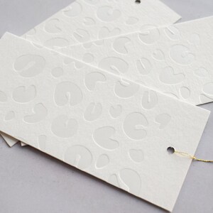 May include: White gift tags with a raised embossed pattern of abstract shapes. The tags have a hole punched in the top and a gold string attached.