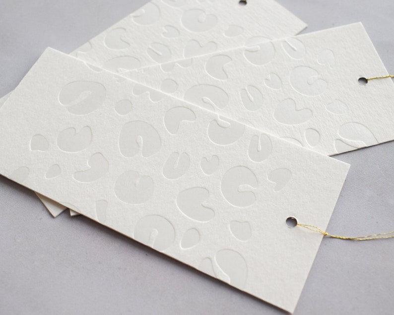 May include: White gift tags with a raised embossed pattern of abstract shapes. The tags have a hole punched in the top and a gold string attached.
