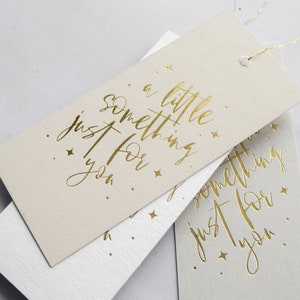 May include: Two beige gift tags with gold foil lettering. The top tag reads "a little something just for you." The bottom tag reads "something just for you."