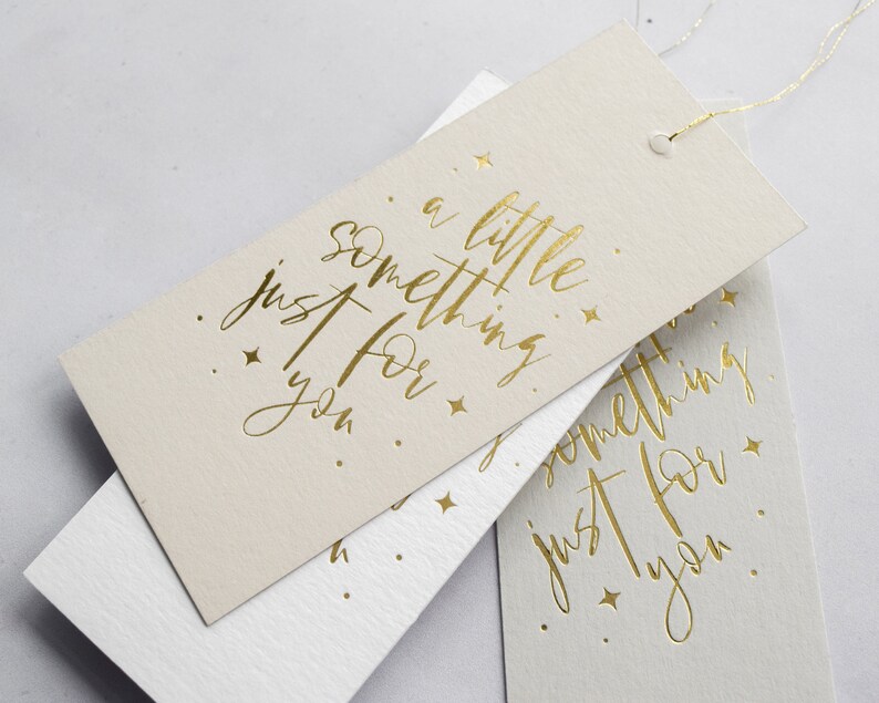 May include: Two beige gift tags with gold foil lettering. The top tag reads "a little something just for you." The bottom tag reads "something just for you."