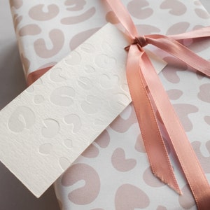 May include: A white gift box with a pink and white leopard print design. The box is tied with a pink satin ribbon and has a blank white gift tag with a similar leopard print design.