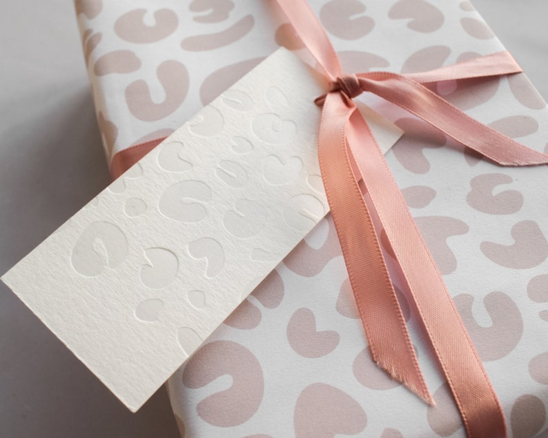 May include: A white gift box with a pink and white leopard print design. The box is tied with a pink satin ribbon and has a blank white gift tag with a similar leopard print design.