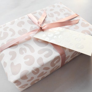 May include: A white gift box with a pink leopard print design. The box is tied with a pink satin ribbon and has a white gift tag attached.