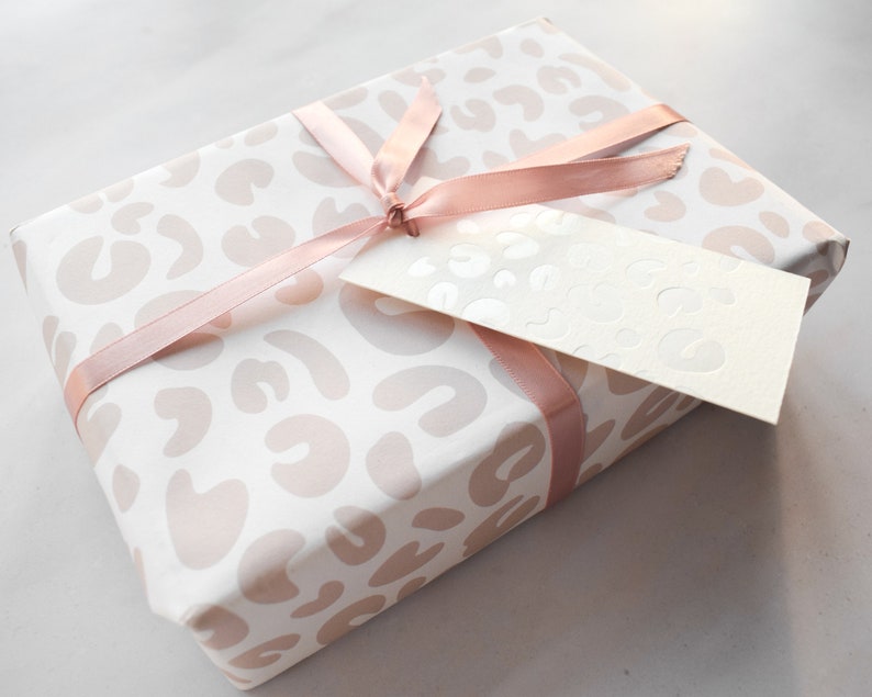 May include: A white gift box with a pink leopard print design. The box is tied with a pink satin ribbon and has a white gift tag attached.