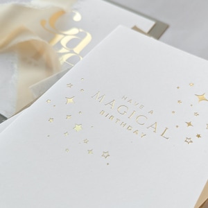 May include: A white card with gold foil lettering that says "Have a Magical Birthday" with a design of gold stars.
