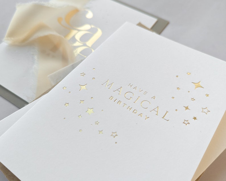 May include: A white card with gold foil lettering that says "Have a Magical Birthday" with a design of gold stars.