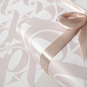 May include: A white gift box with a pink pattern of stylised letters is wrapped with a blush pink satin ribbon tied in a bow.
