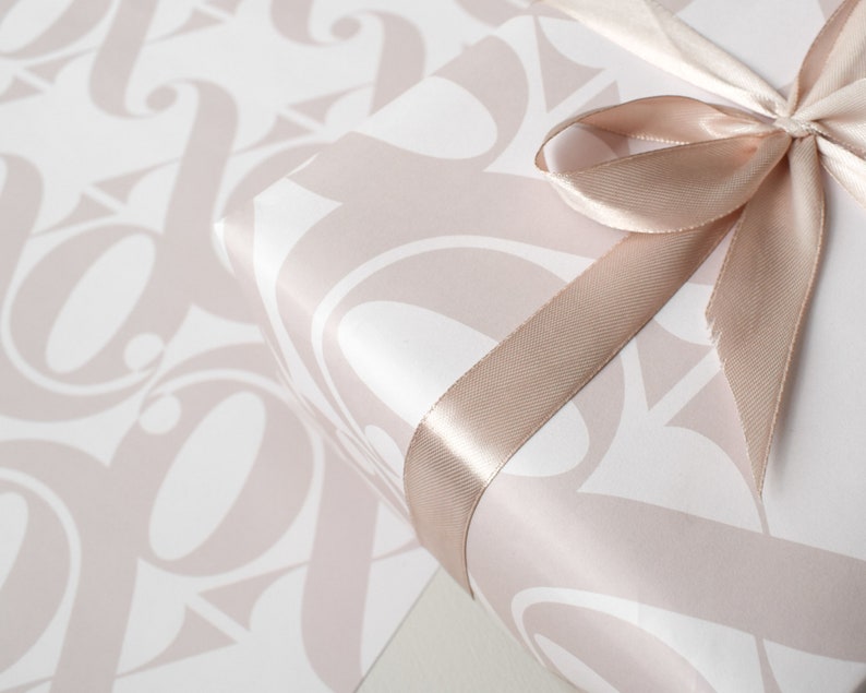 May include: A white gift box with a pink pattern of stylised letters is wrapped with a blush pink satin ribbon tied in a bow.