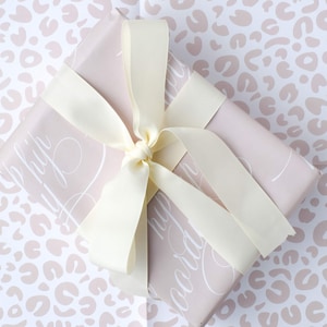 May include: A light pink gift box with a white leopard print pattern. The box is tied with a white satin ribbon and has the words "Happy Birthday" written on it in white script.