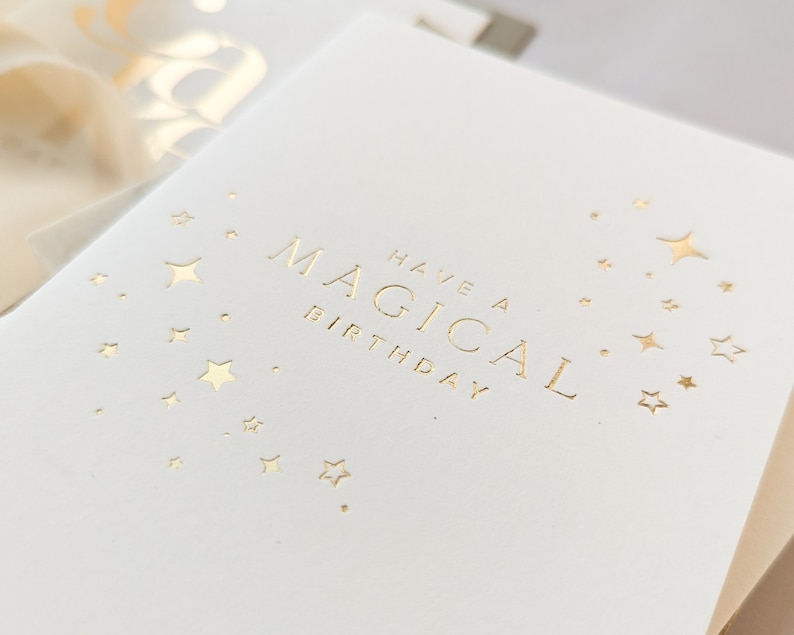 May include: A white card with gold foil lettering that says "Have a Magical Birthday" with gold foil stars scattered around the text.