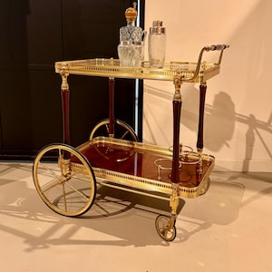 Könnte beinhalten: Goldfarbener Servierwagen mit rechteckiger Ober- und unterer Ablage, beide mit Mahagoni-Finish. Der Wagen hat goldene Räder und Akzente, mit einem Griff und dunklen Holzdetails. Er enthält eine Karaffe, einen Eiskübel und einen Cocktailshaker.